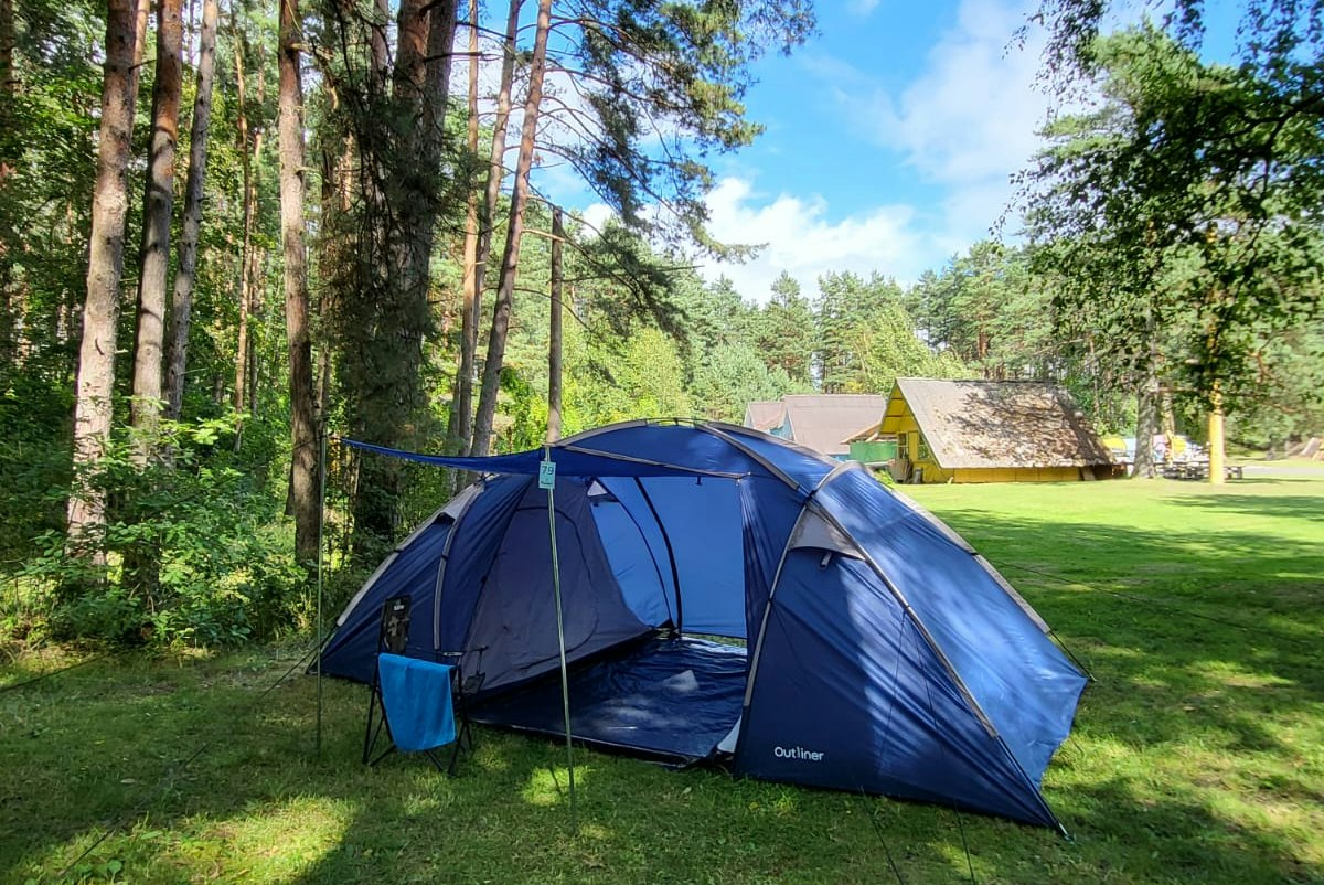 Blaues Campingzelt auf einer grünen Wiese im Wald, mit weiteren Zelten und einem Weg zwischen den Bäumen.