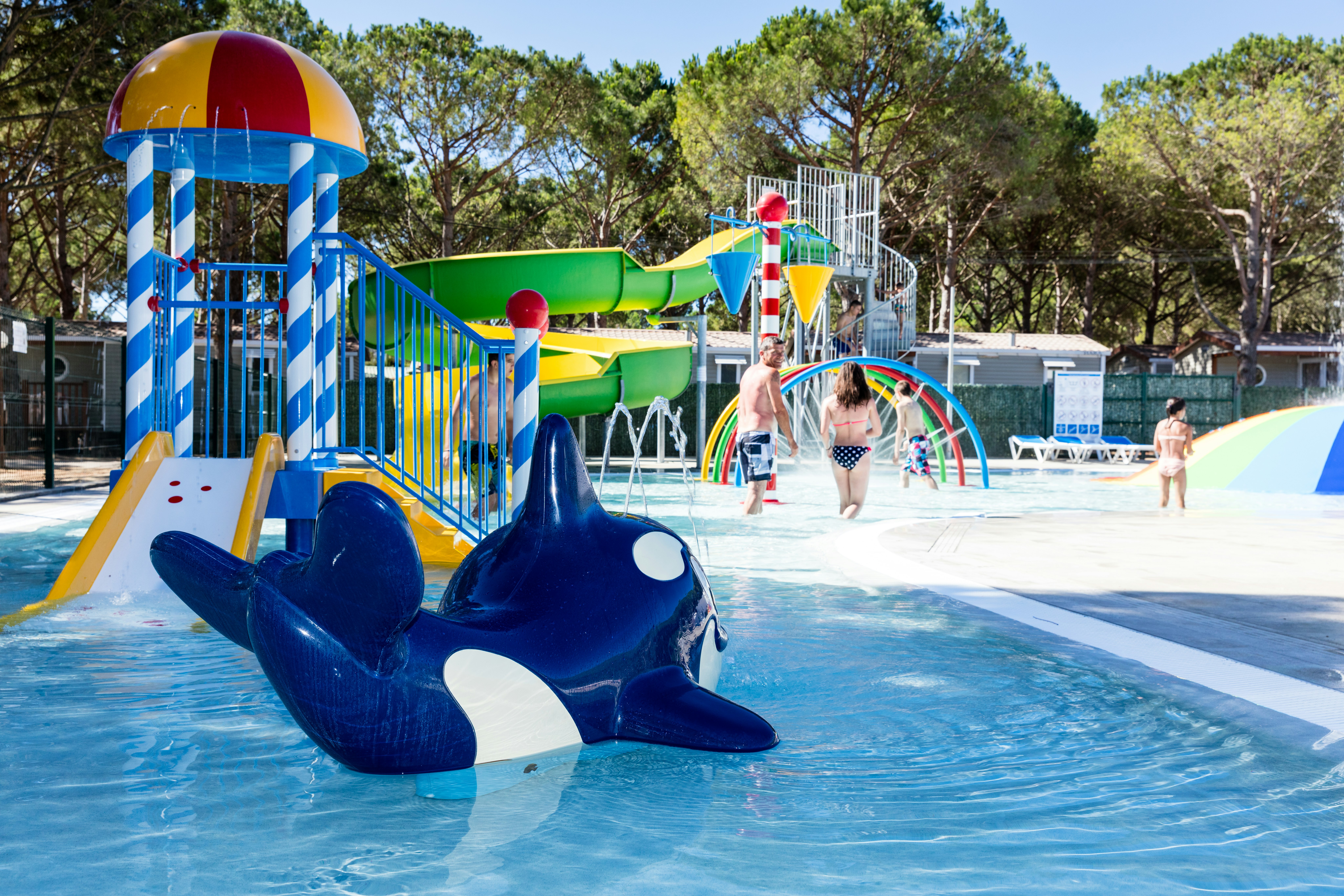 Wasserspielplatz im Pool mit Rutsche, aufblasbarem Wal und bunten Spritzfiguren vor Bäumen.