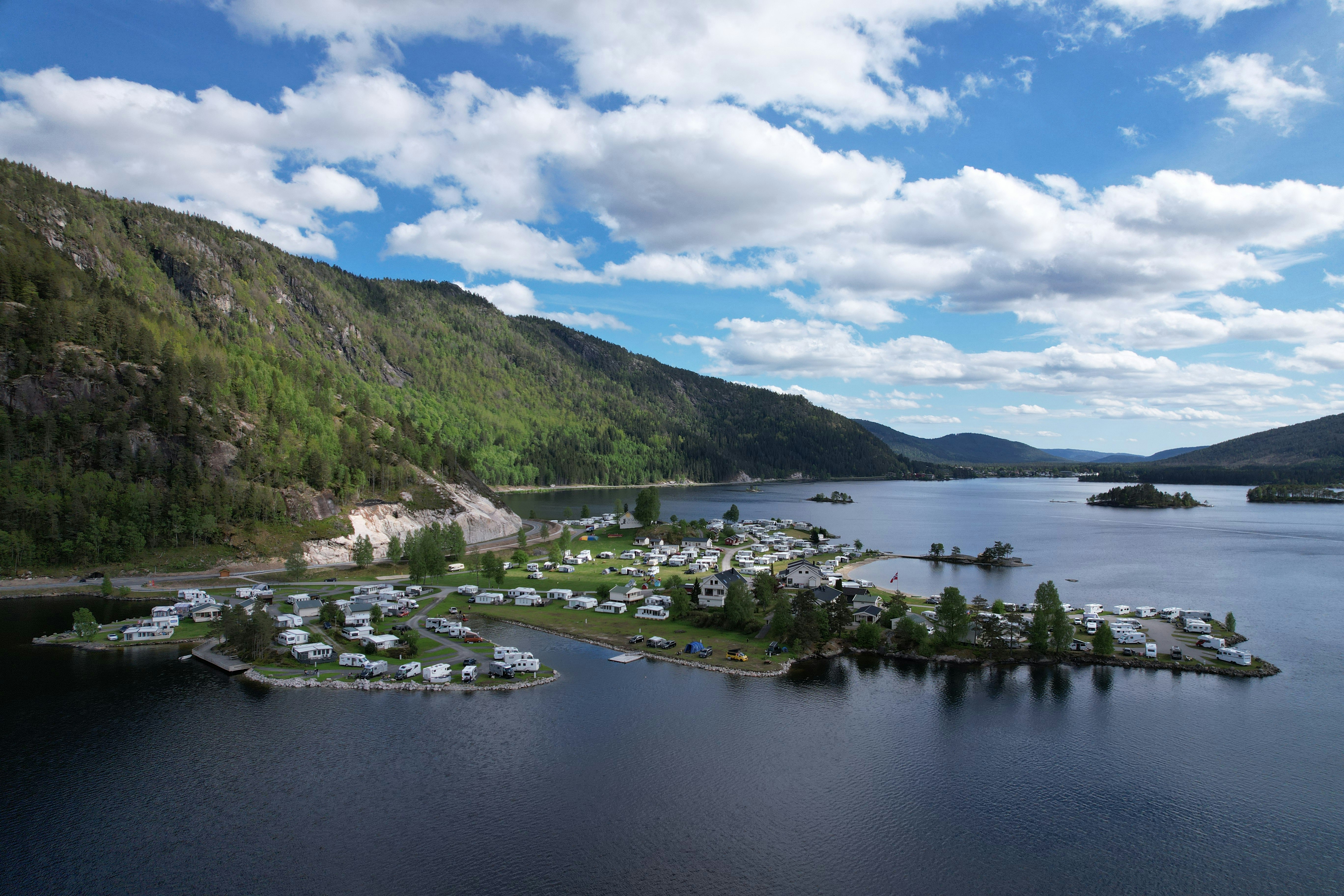 Blick auf einen Campingplatz am See mit vielen Stellplätzen, Booten und kleinen Gebäuden am Ufer, umgeben von Bergen und Bäumen.