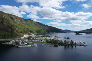 Blick auf einen Campingplatz am See mit vielen Stellplätzen, Booten und kleinen Gebäuden am Ufer, umgeben von Bergen und Bäumen.