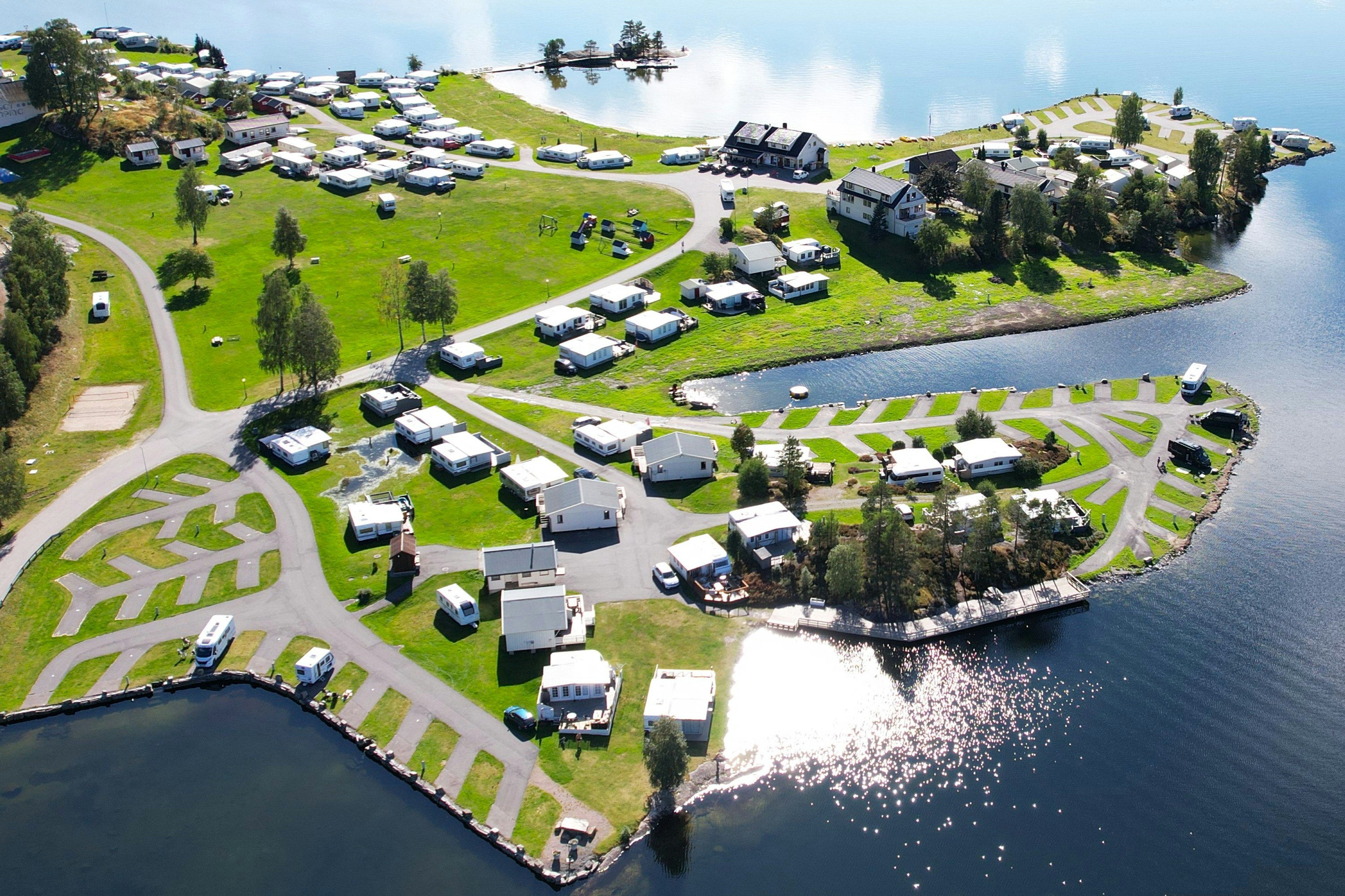 Campingplatz am See mit vielen Stellplätzen und Wohnwagen, umgeben von Grün und Bäumen, Blick von oben auf das Ufer.