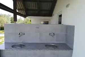Außenküche am Campingplatz mit zwei Spülbecken, Wasserhähnen und grauen Fliesen unter einem überdachten Bereich.