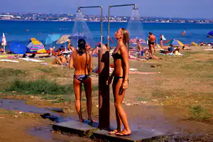 Menschen stehen an einer Außendusche am Strand, im Hintergrund Meer, Liegestühle und Sonnenschirme.