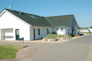 Rezeption eines Campingplatzes: weißes Gebäude mit Satteldach, Eingangstür, Weg und Terrasse mit Steinumrandung.
