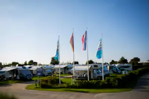 Campingplatz mit Wohnwagen und mehreren Flaggen auf hohen Masten, dazwischen Rasenflächen und ein breiter Weg.