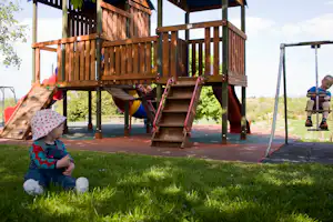 Holz-Spielplatz mit Kletterturm, Rutsche und Schaukel auf gepflasterter Fläche, im Hintergrund Wiese und Bäume.
