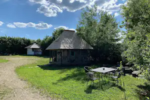 Holzhütte mit kleinem Tisch und Stühlen auf einer grünen Wiese, daneben ein weiteres Gebäude und Bäume.