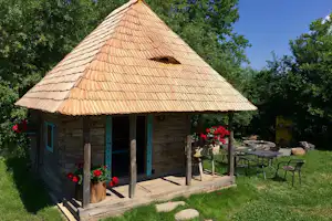 Holzhütte mit Veranda, Ziegeldach und Sitzbank im Garten, umgeben von Bäumen und Rasenfläche.