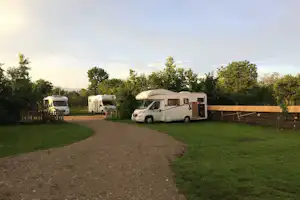 Wohnmobile auf einem Campingplatz auf einem Kiesweg, umgeben von Rasen und Hecken, mit Bäumen im Hintergrund.