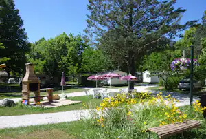 Campingplatz im Grünen mit Blumenbeeten, Sitzbereich unter einer Markise und einem kleinen Holzschild am Weg.