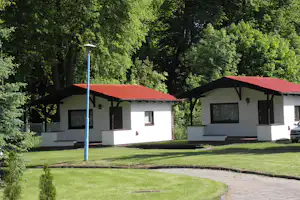 Campinghaus mit rotem Satteldach, weißen Wänden und Fenstern, umgeben von Bäumen und Rasenfläche.