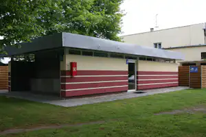 Campinggebäude mit Glasfront, roten Türen und Fensterreihen, davor gepflasterter Weg und Holzsichtschutz, umgeben von Bäumen.