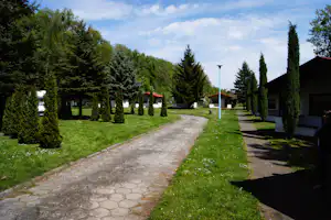 Campingplatz mit gepflastertem Weg, Rasenflächen und Reihen von hohen Zypressen vor mehreren Bungalows mit roten Dächern.
