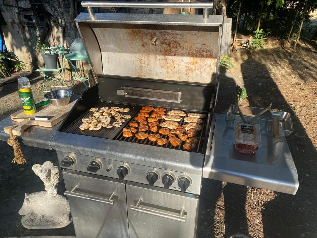 Großer Gasgrill mit Bratgut auf dem Rost, daneben Grillzubehör und Schubladen, im Außenbereich am Campingplatz.