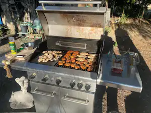 Großer Gasgrill mit Bratgut auf dem Rost, daneben Grillzubehör und Schubladen, im Außenbereich am Campingplatz.