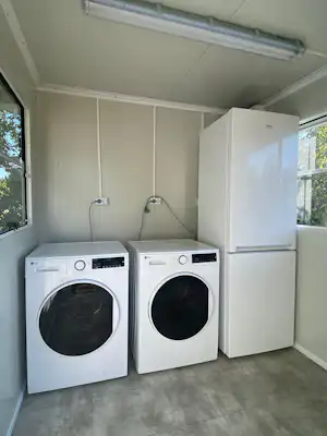 Zwei Waschmaschinen und ein Kühlschrank in einem hellen Raum mit Fliesenboden und Fenstern.