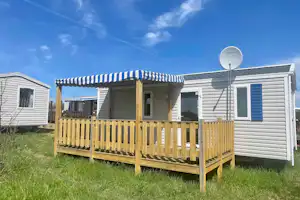 Mobilheim mit Holzveranda und Markise, daneben weitere Wohnwagen auf grünem Campingplatz.