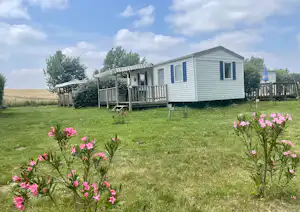 Wohnwagen auf einem Campingplatz mit Holzterrasse, Blumenbeeten im Vordergrund und Rasenfläche vor dem Gebäude.