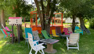 Bunte Campingstühle und ein Tisch auf einer grünen Wiese, im Hintergrund ein Spielbereich mit orangefarbenen Rahmen.