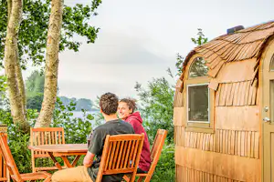 Holzfass-Campingunterkunft mit Fenster und Sitzgruppe; zwei Personen sitzen an Holzbänken vor dem Gebäude im Grünen.