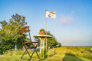 Campingstuhl und Tisch mit Flagge auf einem weißen Säulenständer, umgeben von Wiese, Bäumen und blauem Himmel.