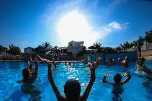 Schwimmbecken im Campingurlaub mit mehreren Personen im Wasser, Sonnenschein, Palmen und Gebäuden im Hintergrund.