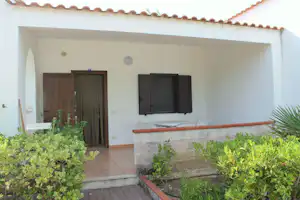 Weiße Ferienhaus-Terrasse mit gepflastertem Boden, Steingrill/Bar und Fensterläden, davor Büsche und ein Holzeingang