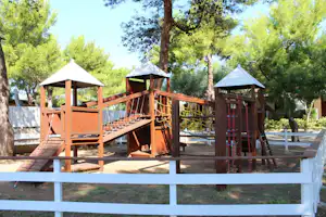 Holz-Spielplatz mit Klettergerüst und Rutsche auf einem Campingplatz, umgeben von Bäumen und einem weißen Zaun.