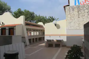 Camping-Terrasse mit zwei Waschbecken, Steinmauer und weißen Wänden, im Hintergrund Gebäude und Bäume.