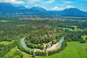 Luftaufnahme eines Campingplatzes am Fluss im grünen Tal, umgeben von Wiesen, Bäumen und Bergen in der Ferne.