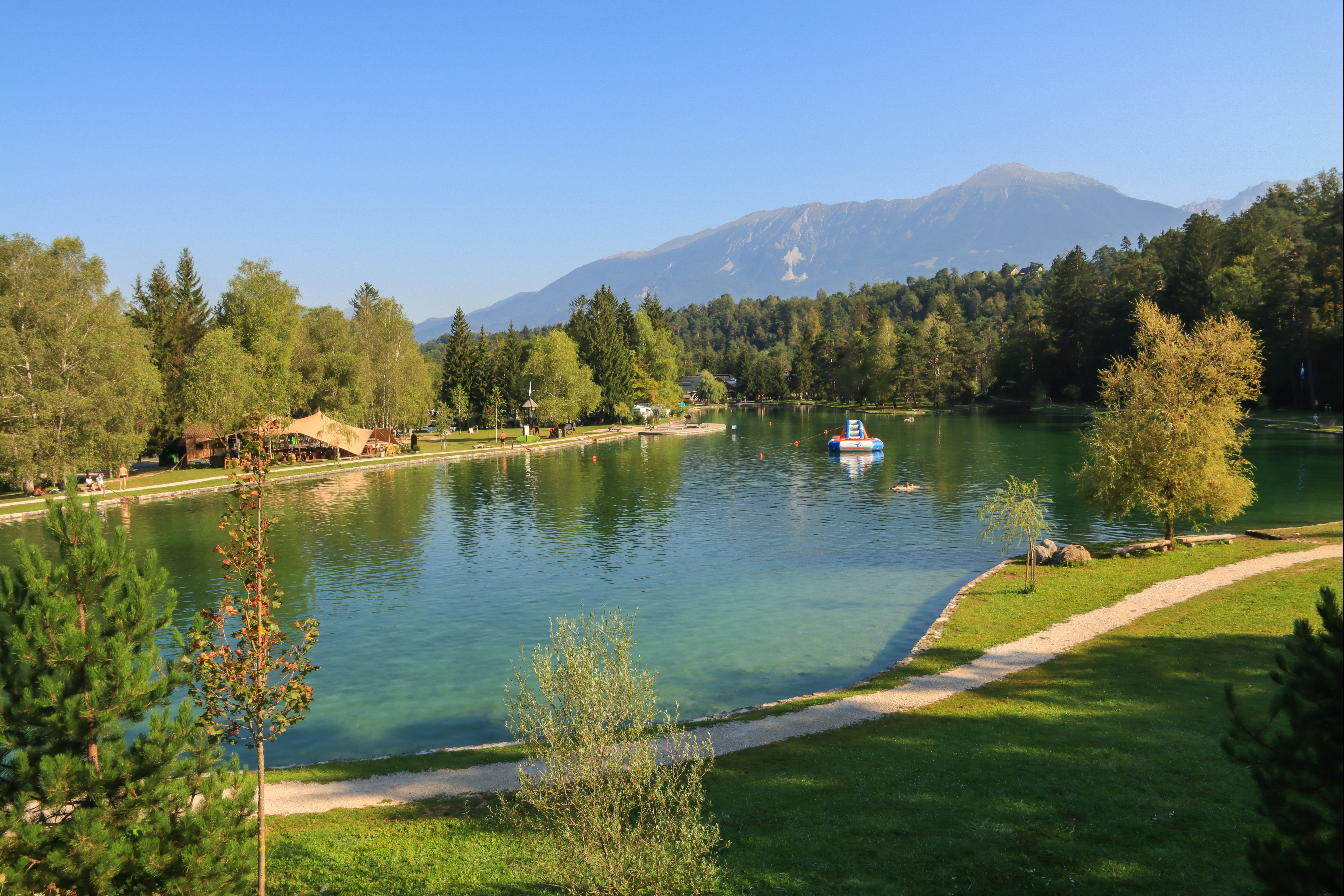 Seeufer am Campingplatz mit Rasen, Kiesweg und Boot auf dem Wasser, im Hintergrund Bäume und Berge.