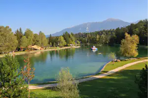 Seeufer am Campingplatz mit Rasen, Kiesweg und Boot auf dem Wasser, im Hintergrund Bäume und Berge.