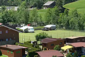 Campingplatz mit Wohnmobilen auf dem Rasen, umgeben von Hecken, Bungalows und einem Tennisplatz im Hintergrund.