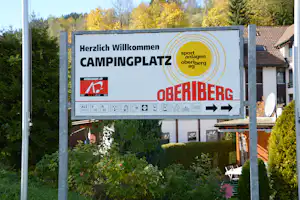 Eingangsschild am Campingplatz Oberiberg mit Beschriftung, Sport- und Berg-Logo, Hecken und Gebäuden im Hintergrund.