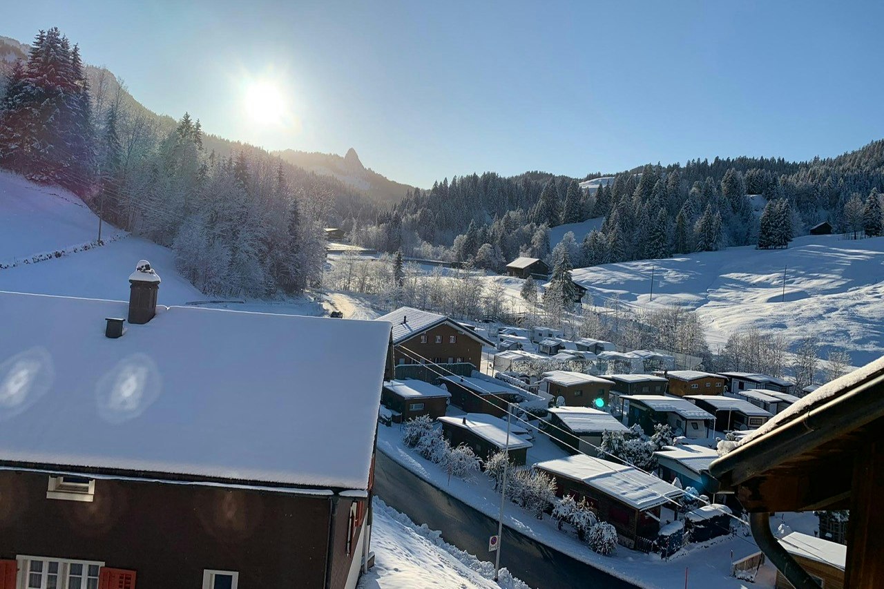 Blick auf verschneite Campinghütten und Wohnwagen in einem Tal, umgeben von Tannen und Bergen bei tiefem Winterlicht.
