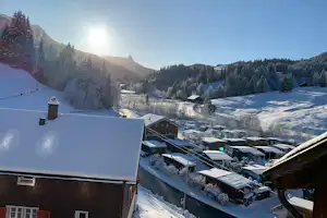 Blick auf verschneite Campinghütten und Wohnwagen in einem Tal, umgeben von Tannen und Bergen bei tiefem Winterlicht.
