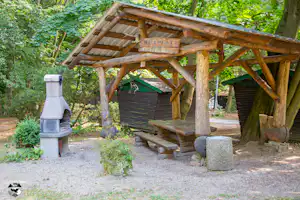 Holzunterstand mit Grillstelle und Sitzbank, davor Steintisch und Feuerstelle, umgeben von Bäumen im Grünen.