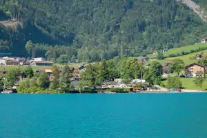 Blick auf den Campingplatz am See mit Wohnhäusern, Wohnmobilen und Bäumen am Ufer vor grünen Bergen.