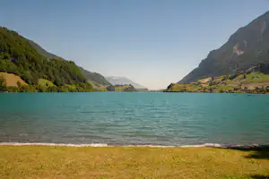 Seeufer mit grünem Gras, Blick auf Berge und bewaldete Hänge, Häuser am Ufer und kleine Boote im Wasser.
