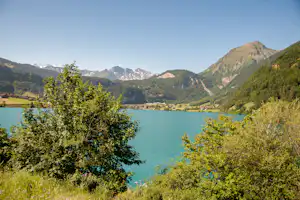 Blick auf einen See mit grünen Bäumen im Vordergrund, dahinter ein Tal mit Häusern und schneebedeckten Bergen.