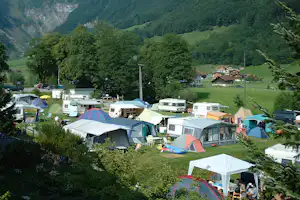 Campingplatz mit vielen Zelten und Wohnwagen auf einer Wiese, umgeben von Bäumen und Bergen im grünen Tal.