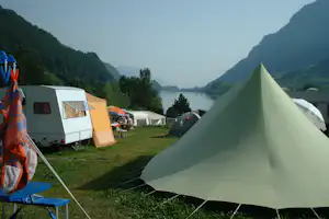 Großes Zelt auf dem Campingplatz, daneben Wohnwagen und weitere Zelte, im Hintergrund ein ruhiger Fluss zwischen Bergen.