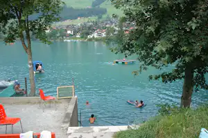 Seeufer mit Badeplattform, Ruderbooten und Blick auf ein Dorf im Tal, eingerahmt von Bäumen.