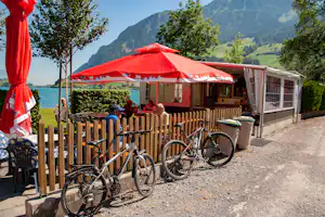 Camping-Terrasse mit roten Sonnenschirmen am See, Holzzaun, Fahrräder und Sitzgruppe vor einem Gebäude mit Vorhängen.