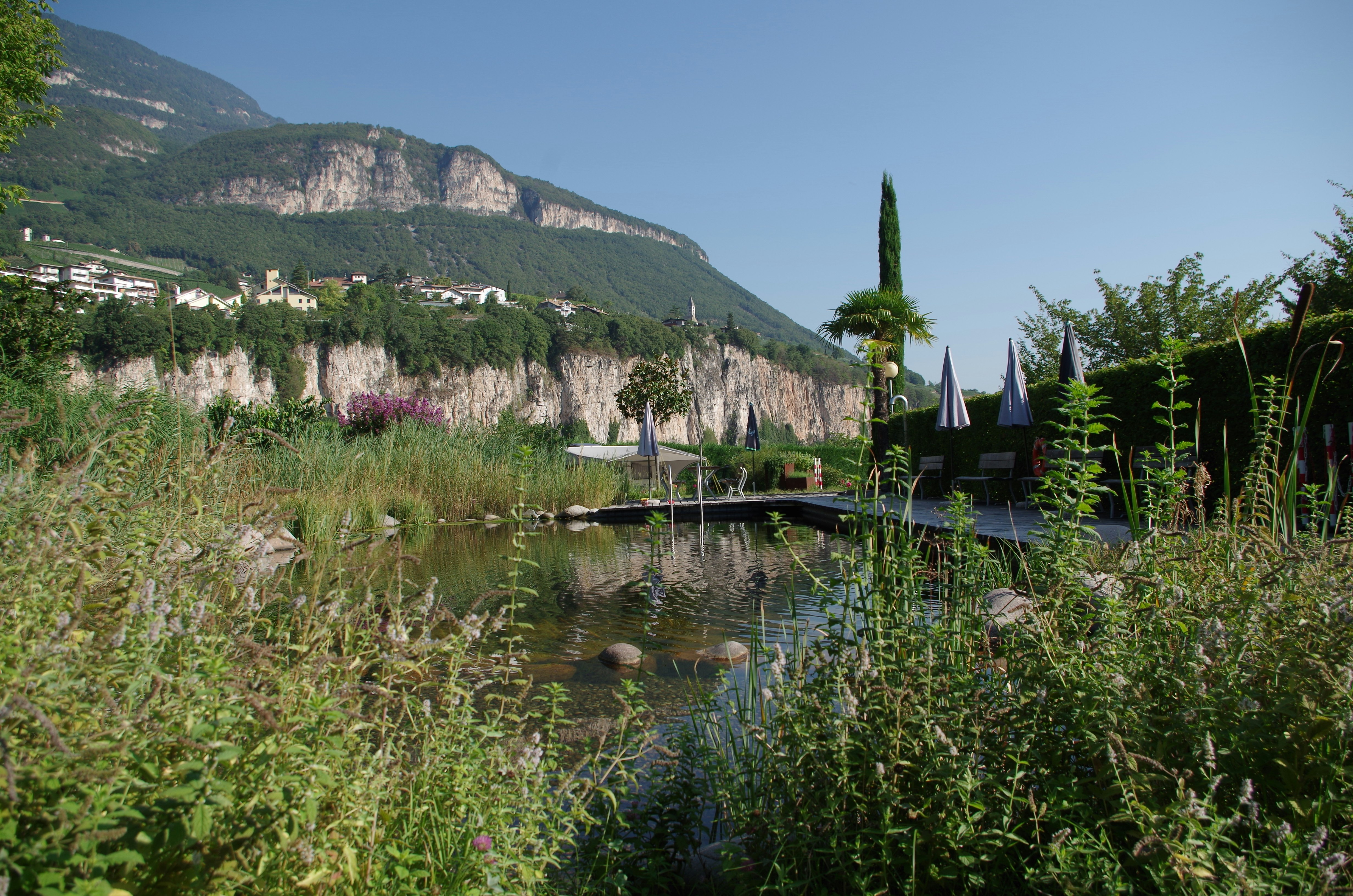 Terrasse am See mit Sonnenschirmen, Liegen und Steg; im Hintergrund Häuser am Hang und Felswand im Tal.