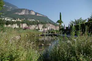 Terrasse am See mit Sonnenschirmen, Liegen und Steg; im Hintergrund Häuser am Hang und Felswand im Tal.