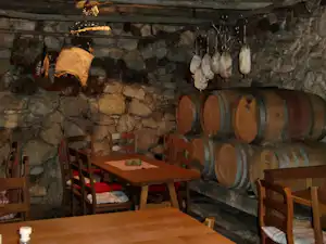 Weinkeller mit Steinmauer, Holzfässern an der Wand und mehreren Holztischen mit Stühlen.