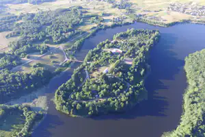 Luftaufnahme eines Campingplatzes am See mit Uferwegen, Bäumen und kleinen Gebäuden inmitten grüner Wiesen.