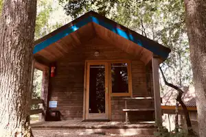 Holzhütte im Wald mit Veranda, Eingangstür und Holztreppe, umgeben von Bäumen und einem kleinen Schild.