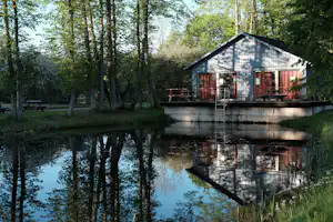 Saunagebäude am Teich im Grünen, mit Holzliegen, Sitzbänken und Leiter am Ufer, umgeben von Bäumen.